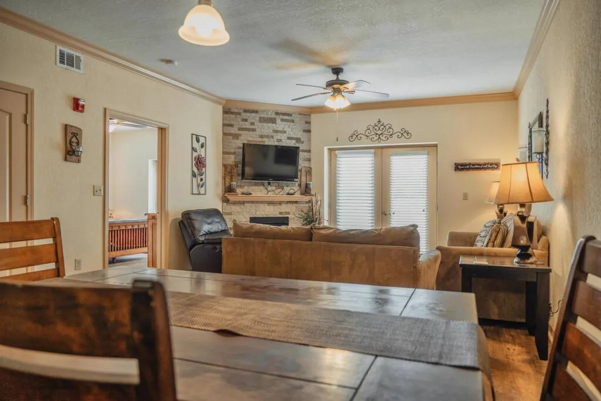 A spacious, inviting open-concept living area featuring a large sofa, dining table, fireplace, and a big flat-screen TV. The scene is arranged for social gatherings, casual dining, and cozy movie nights, with ambient lighting enhancing the atmosphere.