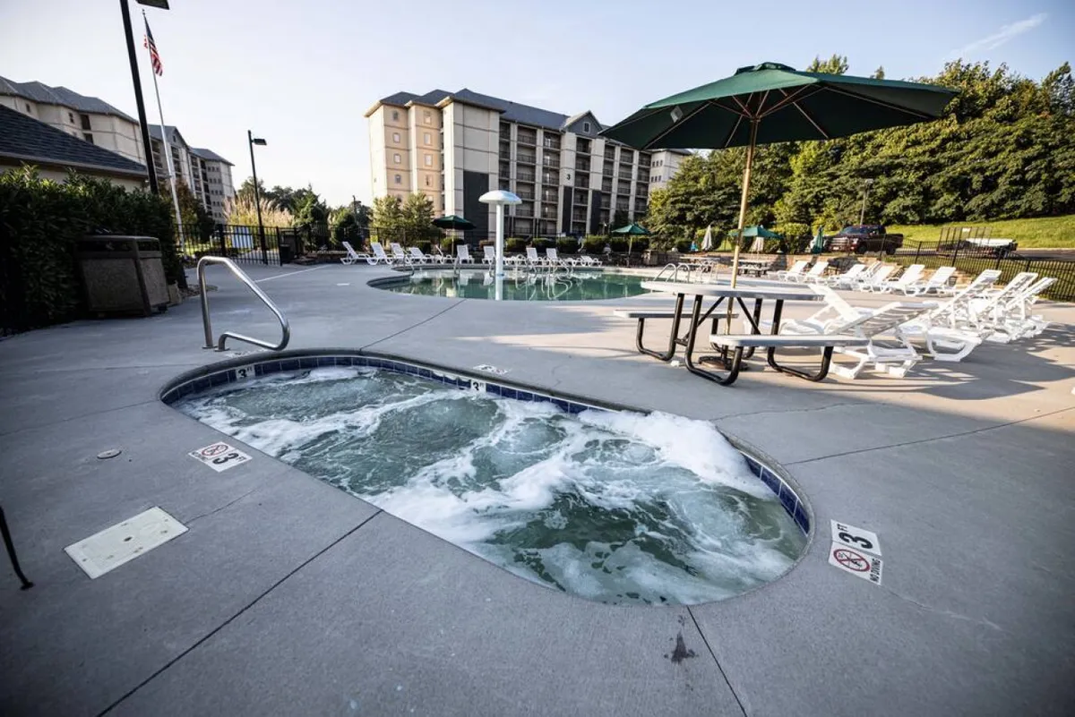 This inviting exterior shows a sparkling swimming pool paired with a hot tub, surrounded by lounge chairs, a picnic table, and an umbrella, all nestled in a relaxed, sunny environment. The space invites socializing, outdoor meals, and relaxation for all ages.