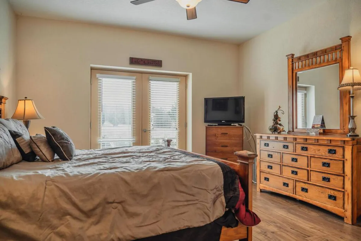 A spacious master bedroom featuring a king-sized bed with decorative pillows, a wooden dresser, large windows with blinds letting in natural light, and a TV mounted on the wall. The overall mood is comfortable and homely, ideal for relaxation and privacy.
