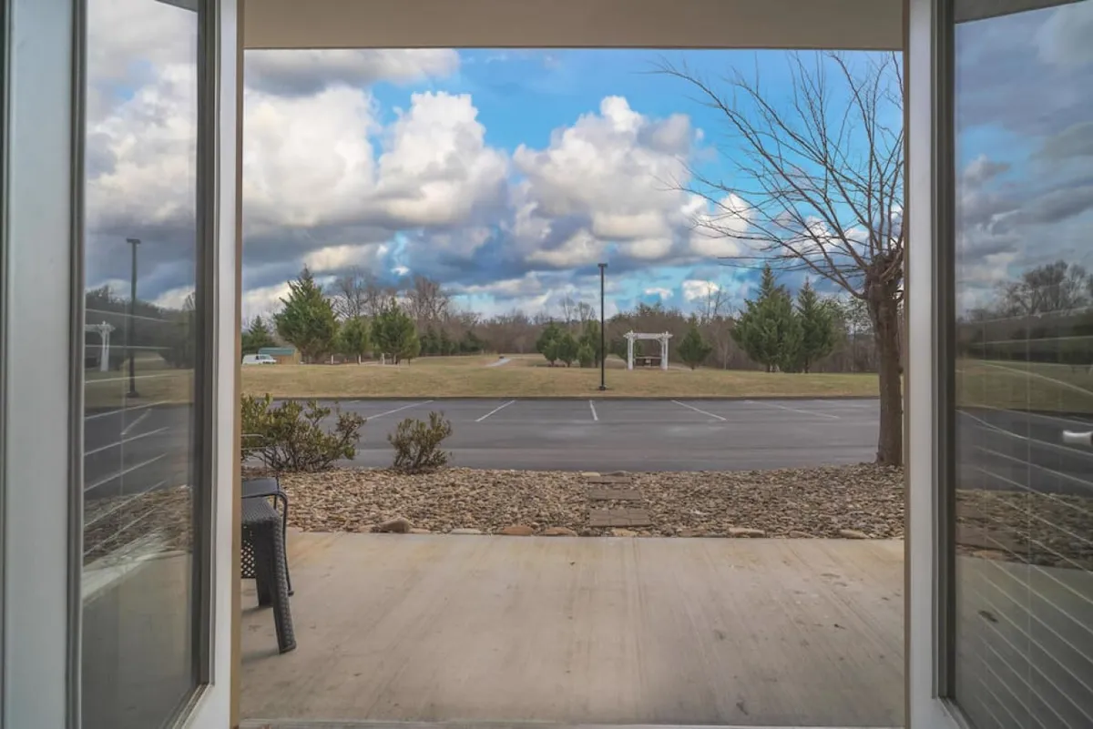 A view from an open doorway looking onto a landscaped area beneath a cloudy sky. The image hints at a tranquil patio or porch space, with inviting seating and lush greenery, perfect for relaxing and enjoying the outdoors. The scene exudes peacefulness and provides a glimpse of the easily accessible, ground-level setting ideal for seniors and couples.