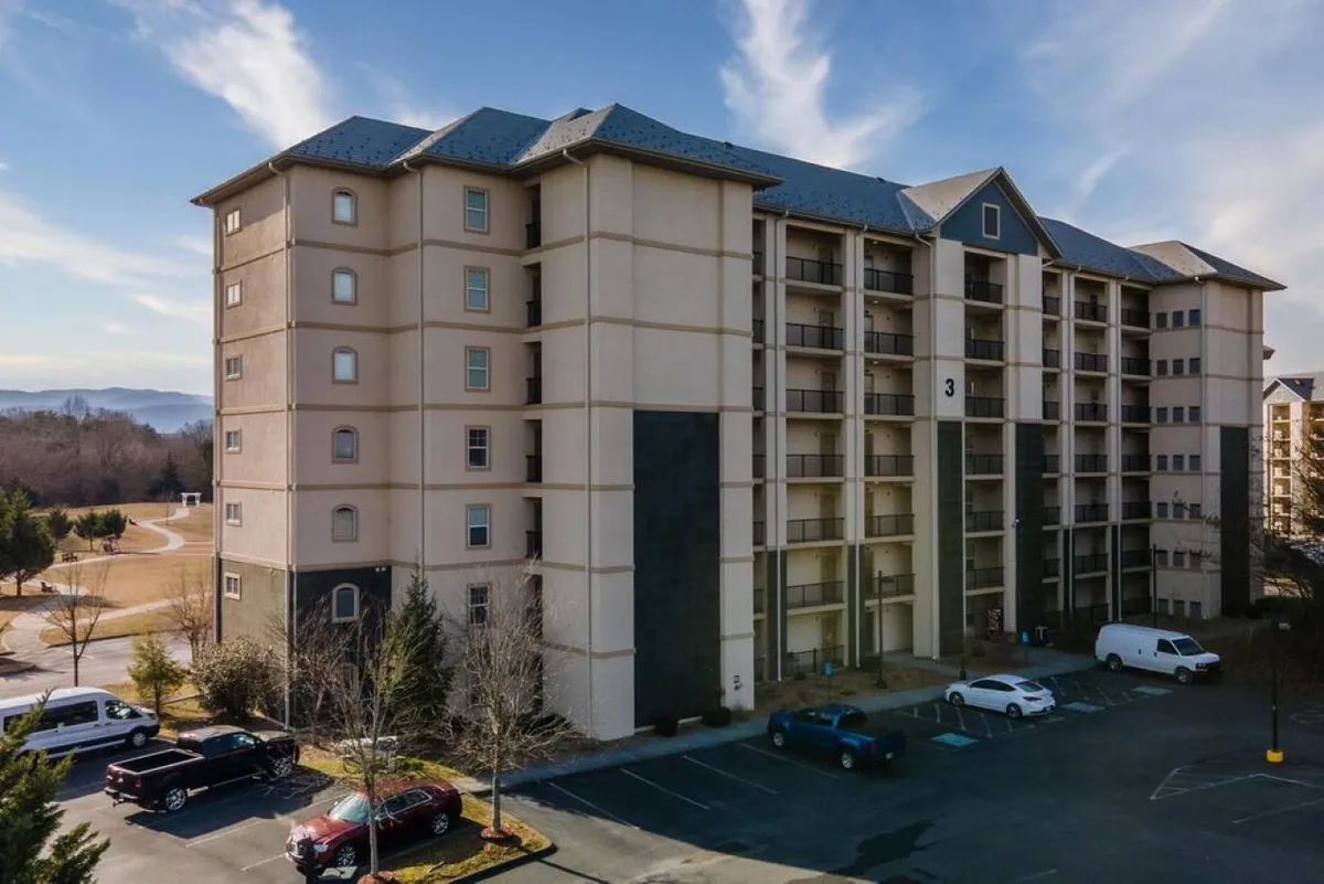 A welcoming view of the multi-story condo building, surrounded by a landscaped area and adjacent parking lot. The scene conveys community living with easy access and a sense of arrival, highlighting accessibility and the building’s inviting presence.
