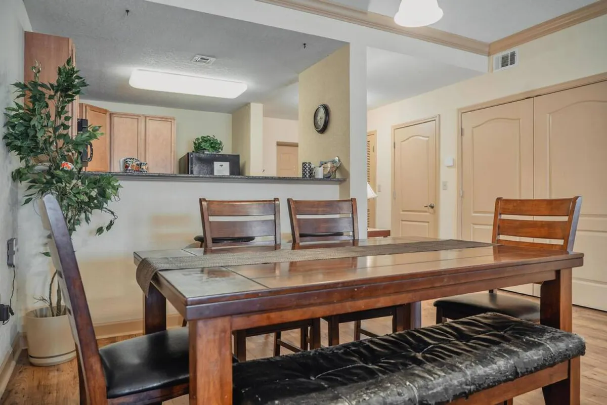 A welcoming dining area with a wooden dining table and chairs, adjacent to a visible kitchen area. A potted plant adds a touch of greenery, and the space is well-lit, suggesting plenty of natural light—ideal for cheerful meals and gatherings.