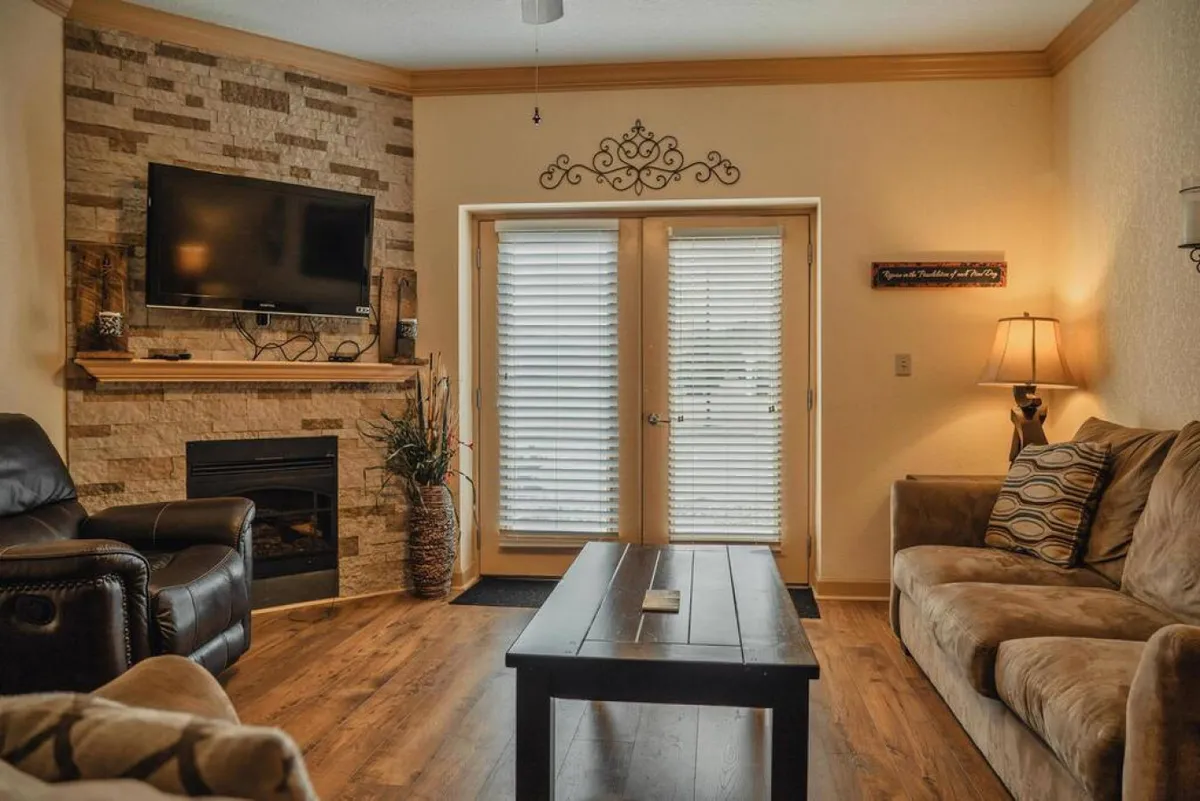A cozy living area with a fireplace, comfortable seating, a wall-mounted TV, and double doors that open to the outdoors. The space is filled with natural light, suggesting easy access to exterior amenities and a seamless connection between indoor and outdoor activities.