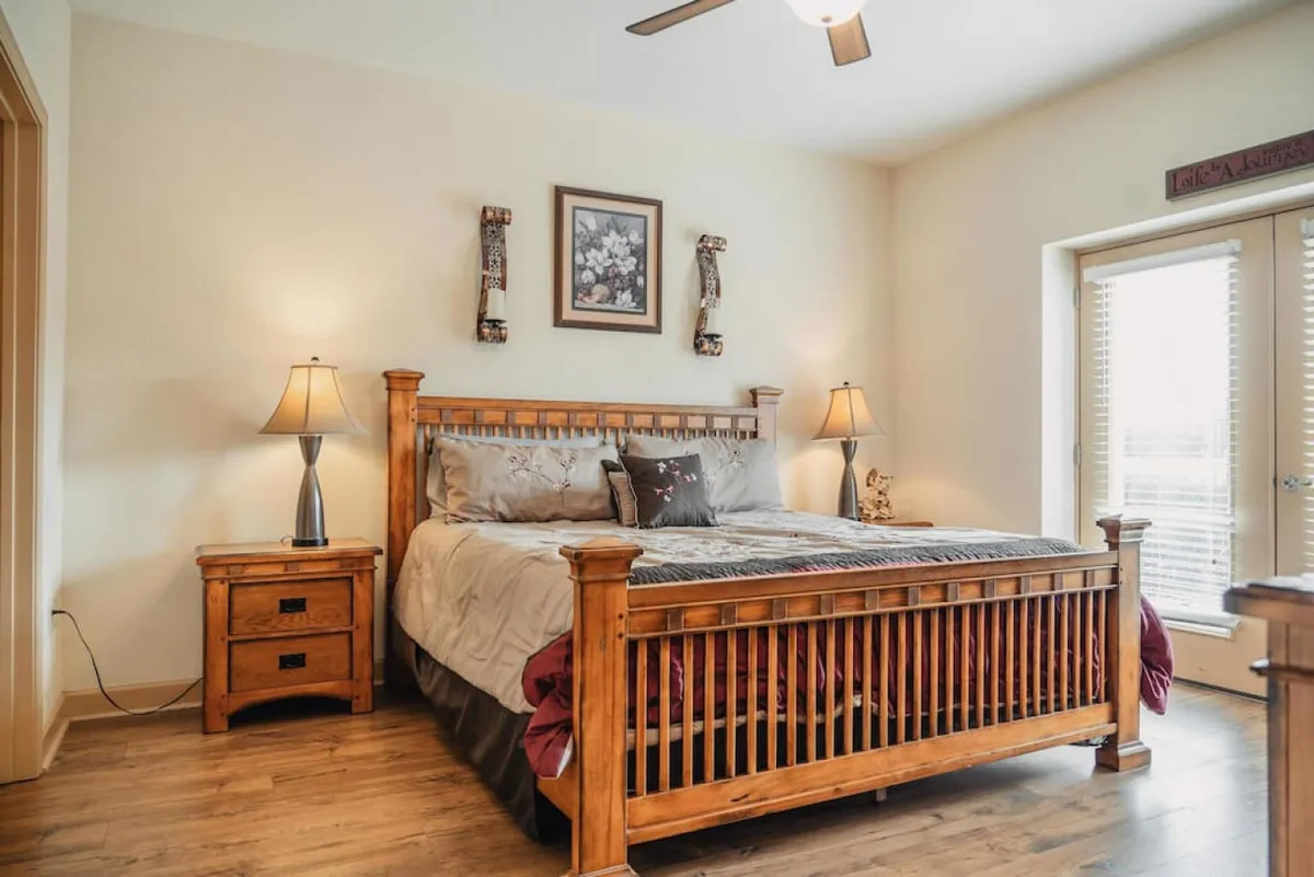 The image showcases a tastefully decorated bedroom with a warm wooden bed frame, inviting linens, and cozy nightstands with softly glowing lamps. Above the bed hangs decorative wall art, while a window floods the space with soothing natural light, creating a tranquil and serene ambiance perfect for unwinding.